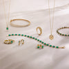A gold jewelry set with vibrant green emerald gemstones, including necklaces, bracelets, rings, and earrings, arranged on a white draped fabric background.