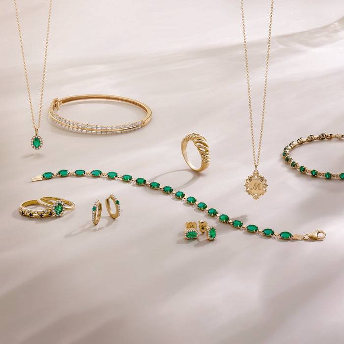 A gold jewelry set with vibrant green emerald gemstones, including necklaces, bracelets, rings, and earrings, arranged on a white draped fabric background.