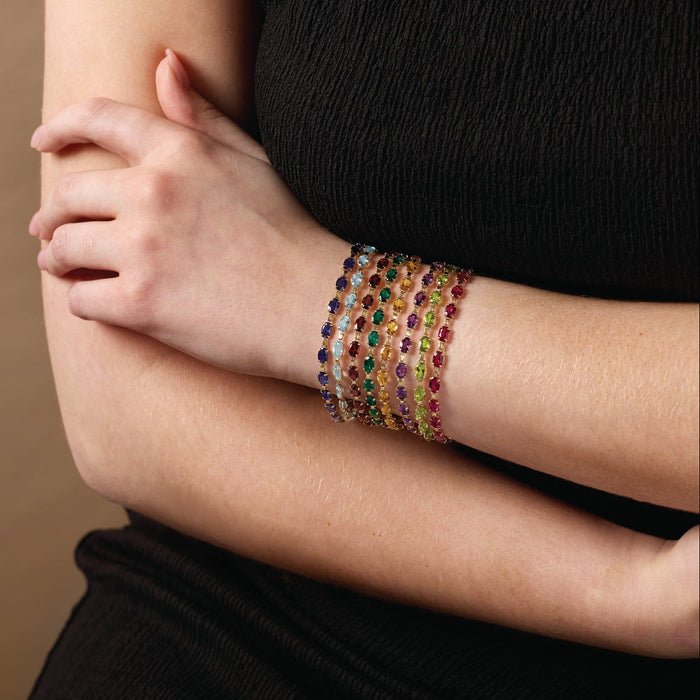 A close-up of a person's arm wearing a stack of seven colorful oval gemstone line bracelets, styled with a black sleeveless top against a warm brown background.