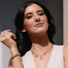 Woman wearing a white sleeveless top and beige pants with jewelry, standing against a dark background.