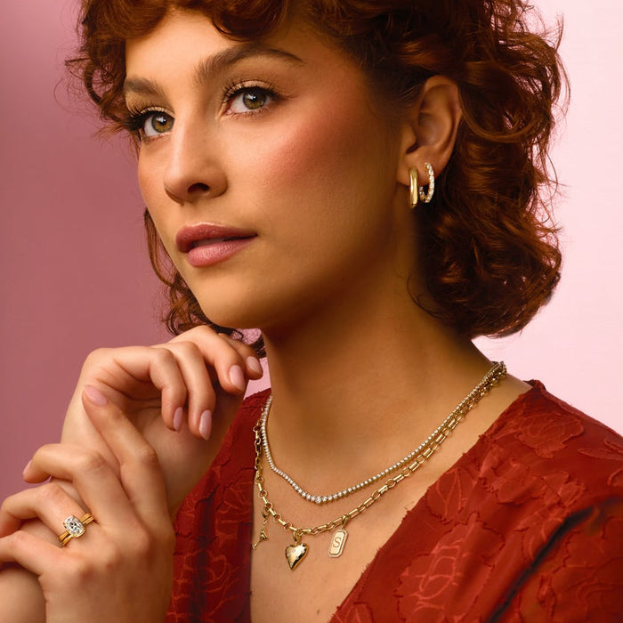 Woman with curly hair and a red top wearing gold jewelry against a pink background