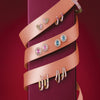 A vertical display of multiple pairs of earrings and a bracelet arranged on a dark burgundy rectangular pedestal, wrapped in a peach satin ribbon. The jewelry features small gold settings with pink enamel and colorful gemstones including pink, blue, and white, set against a solid dark red background.