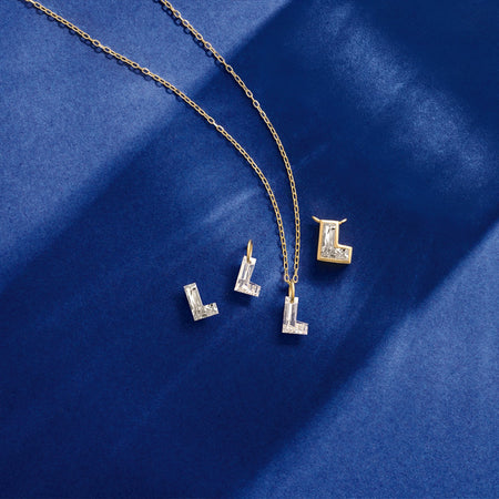 A gold chain necklace with three letter 'L' pendants, each adorned with floating diamond initials, displayed on a textured dark blue fabric background.