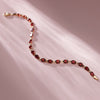 A delicate gold chain bracelet with oval red garnet gemstones, laid out in a curved line on a soft pink background with diagonal light rays.