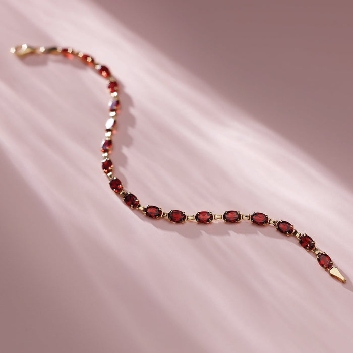A delicate gold chain bracelet with oval red garnet gemstones, laid out in a curved line on a soft pink background with diagonal light rays.