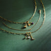Gold layered chain necklaces with whimsical charms, including a star, crescent moon with pearl, and small purple and green accents, displayed on a textured dark green surface with dramatic light rays.
