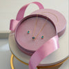 A delicate gold necklace and matching stud earrings with small blue-green gemstones, presented in an open round pink jewelry box with a pink satin ribbon, displayed on a white and gold tiered stand.