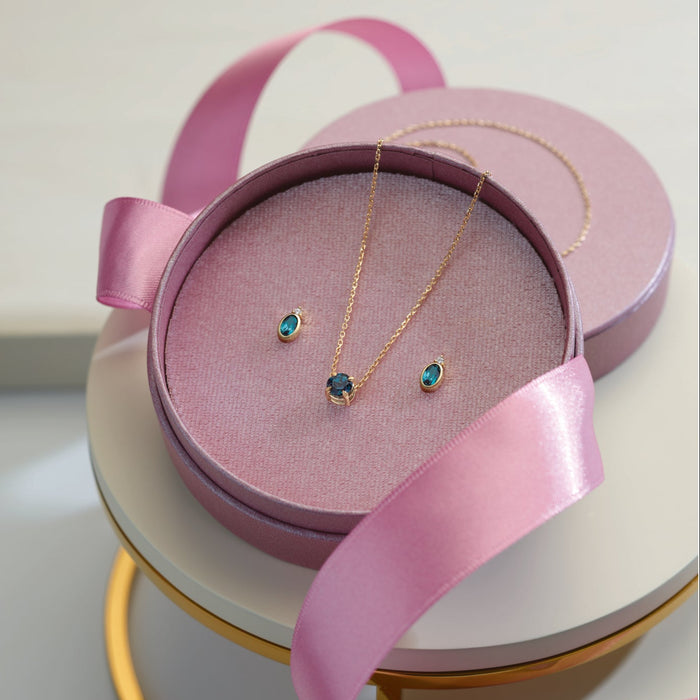 A delicate gold necklace and matching stud earrings with small blue-green gemstones, presented in an open round pink jewelry box with a pink satin ribbon, displayed on a white and gold tiered stand.