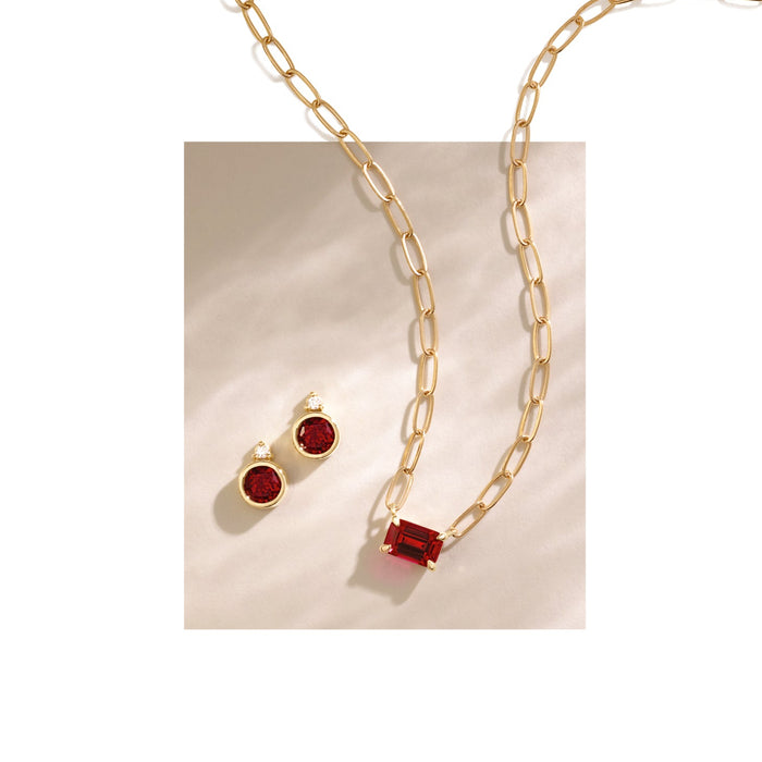 Gold paperclip chain necklace with a rectangular red gemstone pendant, displayed with matching gold stud earrings featuring round red gemstones on a light beige textured background.