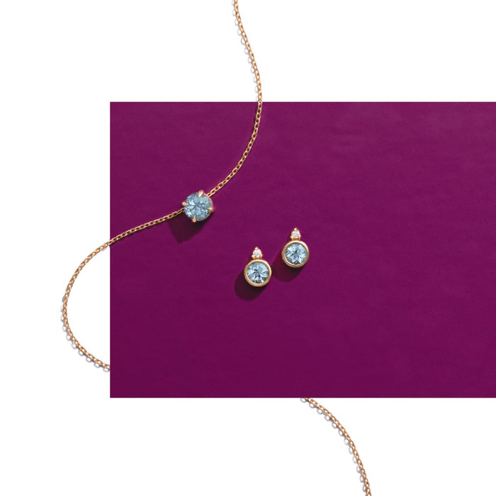 A gold chain necklace and matching stud earrings with round light blue gemstones, arranged on a solid magenta rectangular card against a white background.