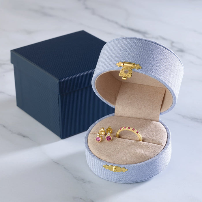 An open round jewelry box with a pale blue exterior, gold-tone clasp, and beige interior, displayed on a white marble surface next to a dark navy gift box. Inside the box are a gold ring and a small gold and pink stud earring.